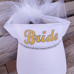 K&ouml;nnte beinhalten: Wei&szlig;e Visier mit einem T&uuml;llschleier und dem Wort "Bride" in Gelb mit Strasssteinen.