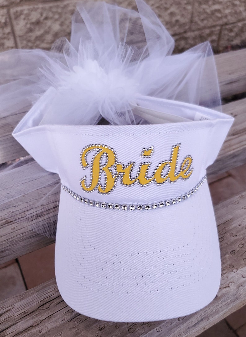 K&ouml;nnte beinhalten: Wei&szlig;e Visier mit einem T&uuml;llschleier und dem Wort "Bride" in Gelb mit Strasssteinen.