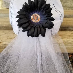 May include: White tulle hat with a black flower and a San Francisco Giants logo. The hat is decorated with a black flower and a small, round, orange and black San Francisco Giants logo.
