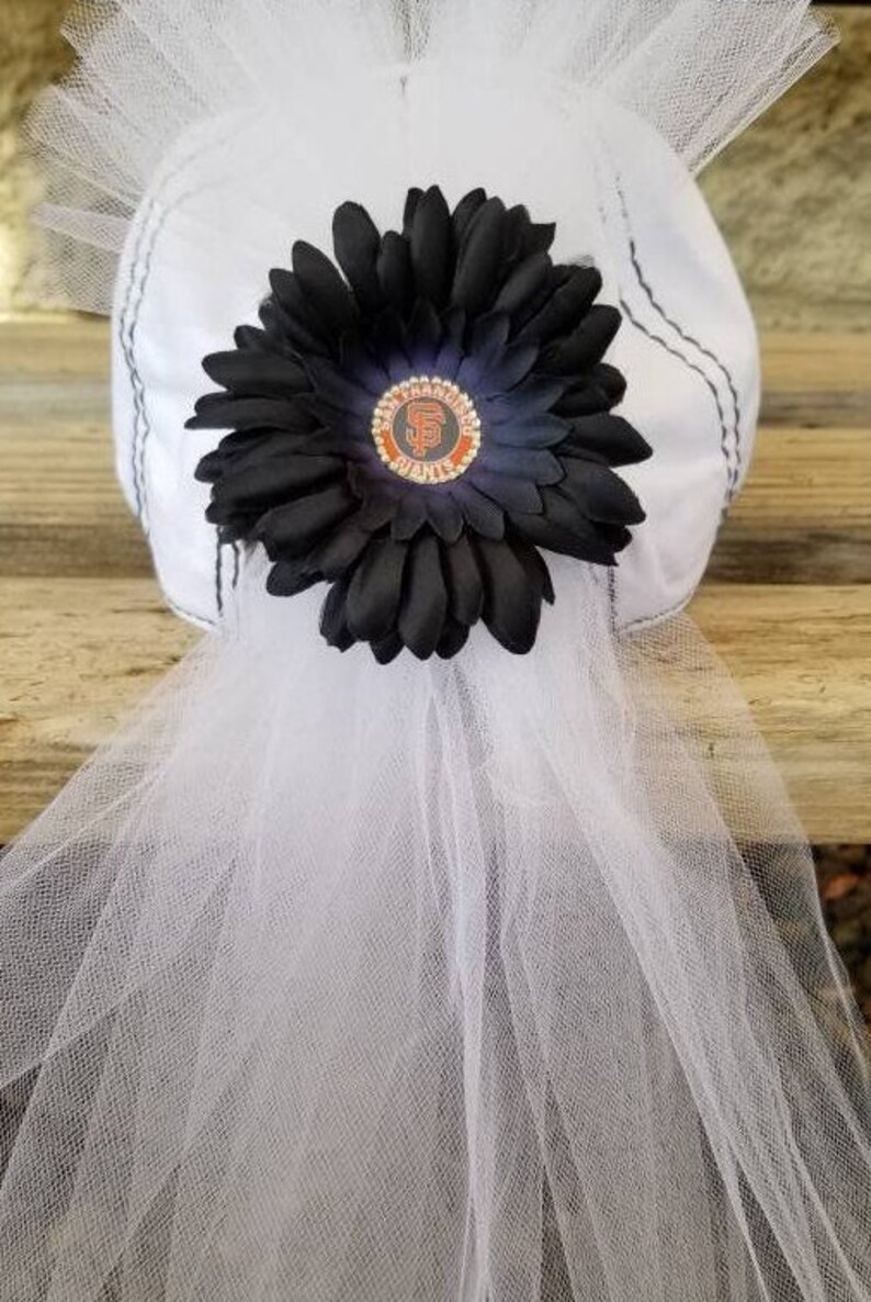 May include: White tulle hat with a black flower and a San Francisco Giants logo. The hat is decorated with a black flower and a small, round, orange and black San Francisco Giants logo.