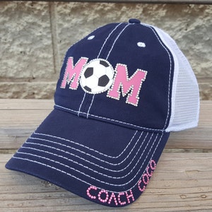 Op de afbeelding: Een marineblauwe en witte baseballpet met een roze "MOM" ontwerp met een voetbal. De pet heeft een mesh achterkant en de woorden "COACH MOM" zijn geborduurd op de klep.