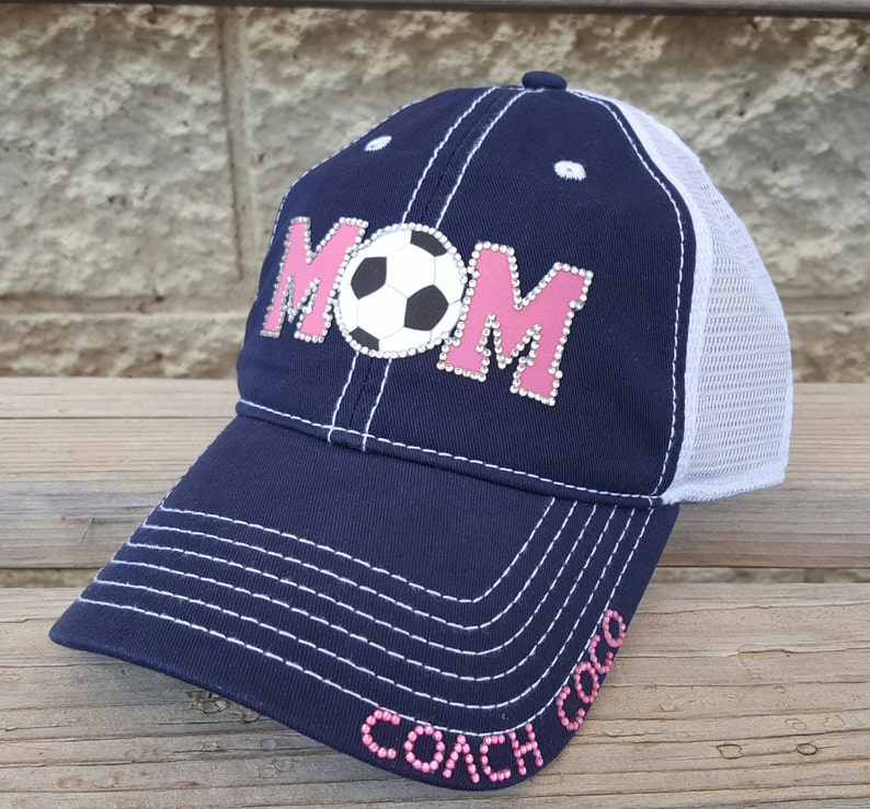 Op de afbeelding: Een marineblauwe en witte baseballpet met een roze "MOM" ontwerp met een voetbal. De pet heeft een mesh achterkant en de woorden "COACH MOM" zijn geborduurd op de klep.