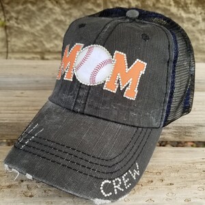 May include: Black and orange baseball cap with a white baseball and the word "MOM" in rhinestones. The cap has a mesh back and the word "CREW" in rhinestones on the brim.