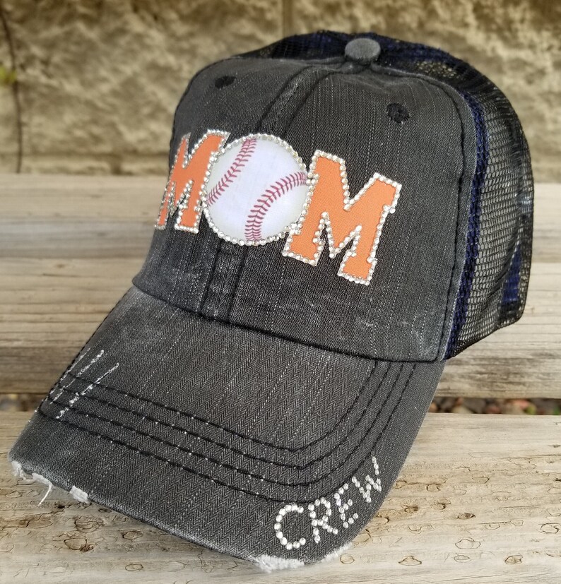 May include: Black and orange baseball cap with a white baseball and the word "MOM" in rhinestones. The cap has a mesh back and the word "CREW" in rhinestones on the brim.