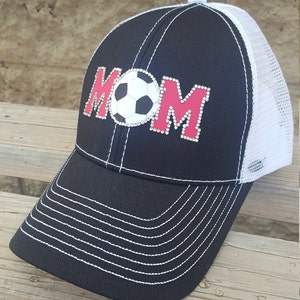 Op de afbeelding: Zwarte en witte truckercap met een rode en witte voetbal en het woord "MOM" in strass.