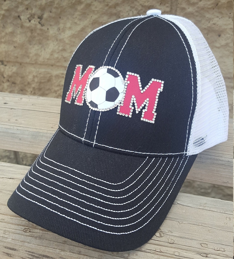 Op de afbeelding: Zwarte en witte truckercap met een rode en witte voetbal en het woord "MOM" in strass.