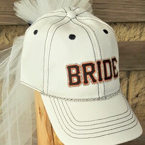 May include: White baseball cap with a black brim and a white tulle veil. The cap has a black and orange "BRIDE" patch with rhinestones.