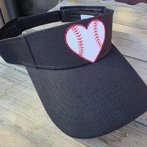May include: A black visor featuring a white heart-shaped patch on the front panel. The heart is designed to resemble a baseball, with red stitching details along the center. The visor has an adjustable strap at the back and a curved brim.