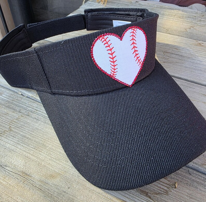 May include: A black visor featuring a white heart-shaped patch on the front panel. The heart is designed to resemble a baseball, with red stitching details along the center. The visor has an adjustable strap at the back and a curved brim.