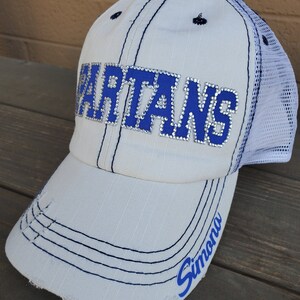 May include: A white baseball cap with a distressed brim and a white mesh back. The front features the word SPARTANS in blue letters outlined with clear rhinestones. The name Simona is printed in blue script on the right side of the brim.