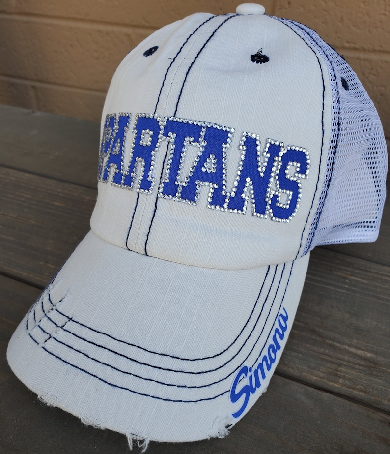 May include: A white baseball cap with a distressed brim and a white mesh back. The front features the word SPARTANS in blue letters outlined with clear rhinestones. The name Simona is printed in blue script on the right side of the brim.