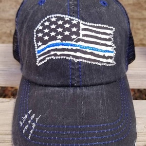 May include: A black distressed denim trucker hat with a mesh back. The hat features a white and blue American flag with a blue line across the middle, made of rhinestones.