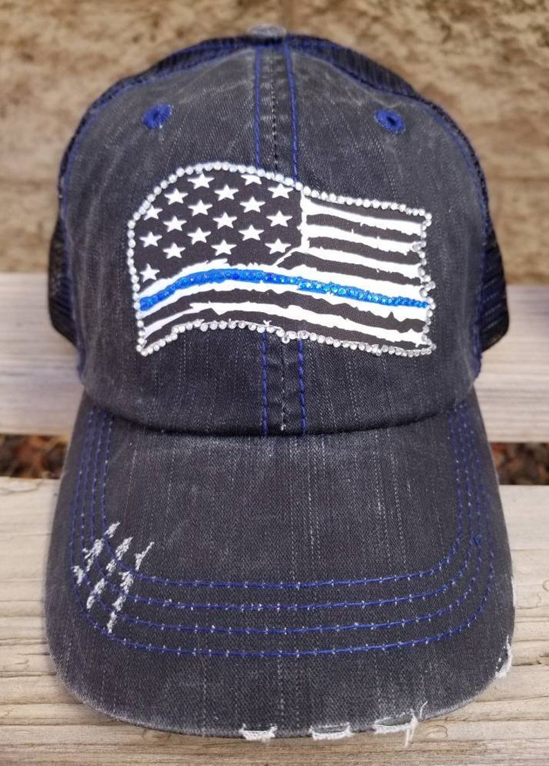 May include: A black distressed denim trucker hat with a mesh back. The hat features a white and blue American flag with a blue line across the middle, made of rhinestones.