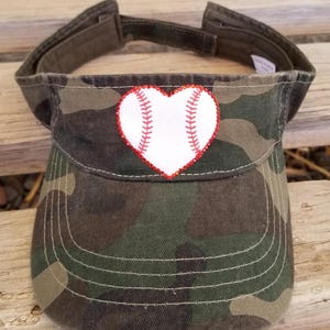 May include: A camouflage print visor featuring a white heart-shaped baseball graphic on the front. The heart is outlined with small red rhinestones and includes red stitching details that mimic the appearance of a baseball's seams.