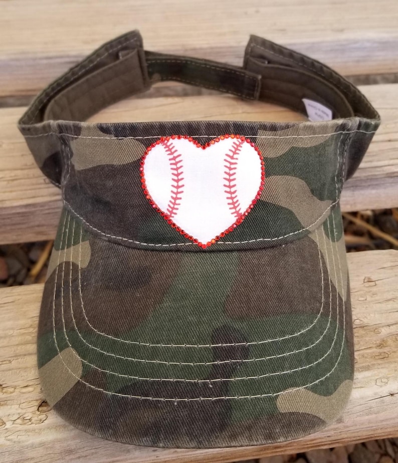 May include: A camouflage print visor featuring a white heart-shaped baseball graphic on the front. The heart is outlined with small red rhinestones and includes red stitching details that mimic the appearance of a baseball's seams.