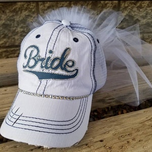May include: White baseball cap with a blue "Bride" design and rhinestones. The cap has a mesh back and a white tulle veil attached to the top.