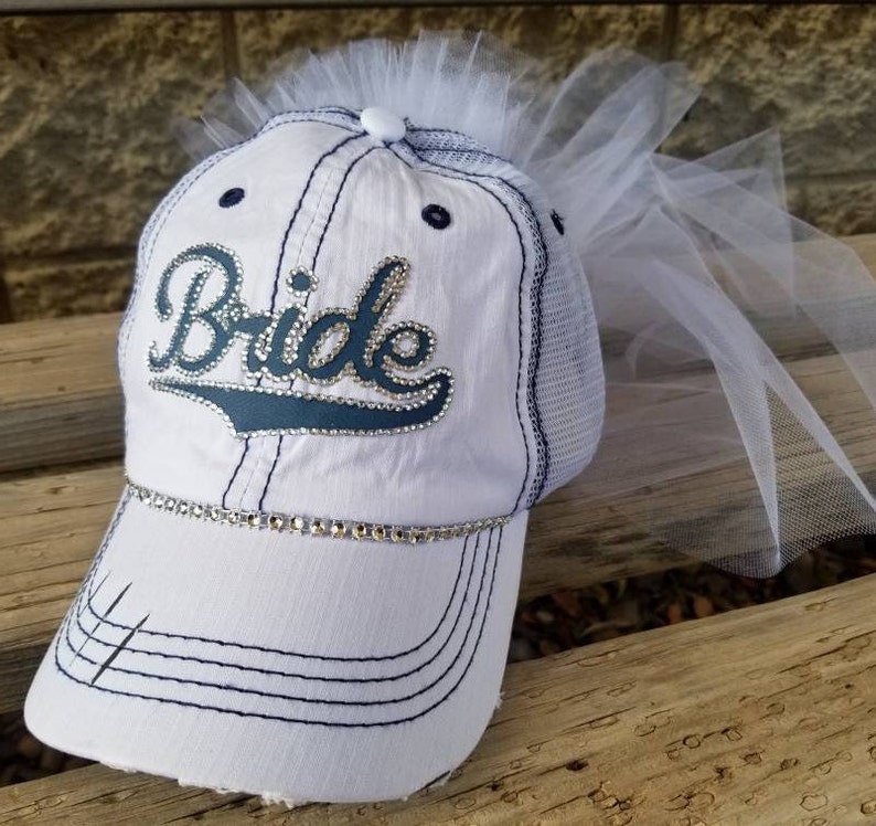 May include: White baseball cap with a blue "Bride" design and rhinestones. The cap has a mesh back and a white tulle veil attached to the top.
