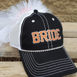 May include: Black and white mesh baseball cap with an orange "BRIDE" design featuring rhinestones. The cap has a white tulle veil attached to the top.