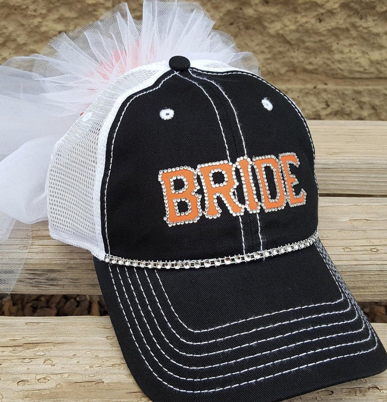 May include: Black and white mesh baseball cap with an orange "BRIDE" design featuring rhinestones. The cap has a white tulle veil attached to the top.