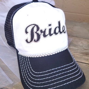 May include: White trucker hat with a black mesh back and a black "Bride" design with rhinestones. The hat has a white tulle veil attached to the top.