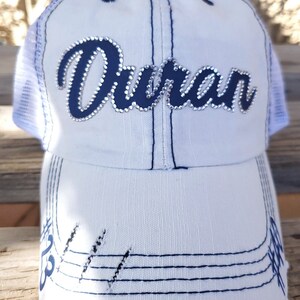May include: White trucker hat with a blue mesh back and a blue cursive word "Duran" with rhinestones on the front. The hat has a distressed look with blue stitching and frayed edges.