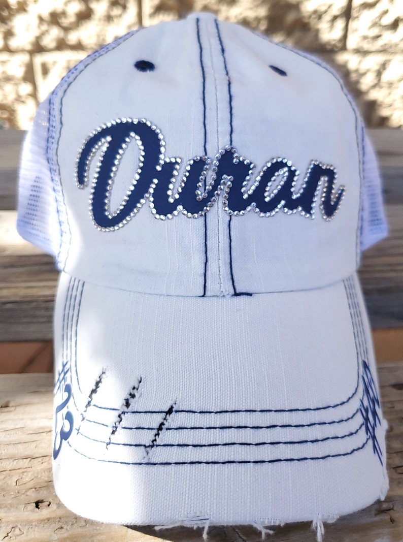 May include: White trucker hat with a blue mesh back and a blue cursive word "Duran" with rhinestones on the front. The hat has a distressed look with blue stitching and frayed edges.