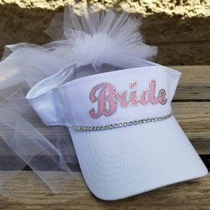 K&ouml;nnte beinhalten: Wei&szlig;e Visier mit einem rosafarbenen "Bride"-Design und Strasssteinen. Die Visier hat einen wei&szlig;en T&uuml;llschleier.
