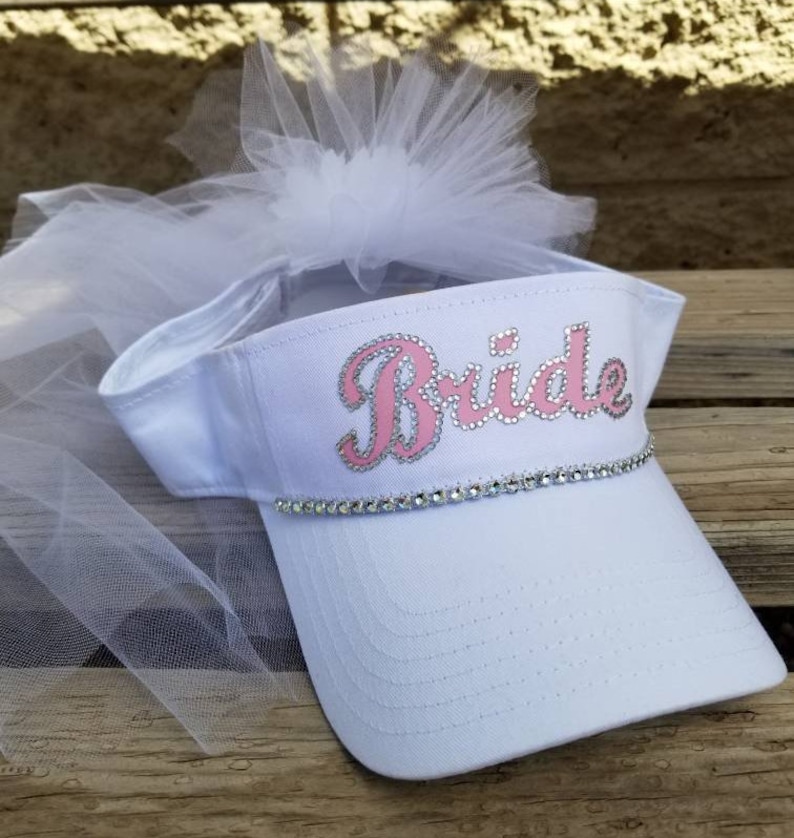 K&ouml;nnte beinhalten: Wei&szlig;e Visier mit einem rosafarbenen "Bride"-Design und Strasssteinen. Die Visier hat einen wei&szlig;en T&uuml;llschleier.