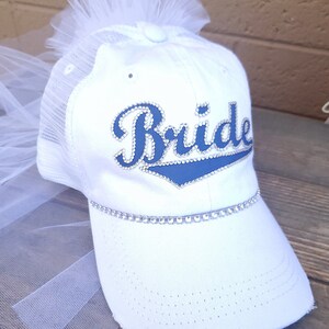 May include: White baseball cap with a tulle veil attached. The cap has a blue and white "Bride" design with rhinestones.