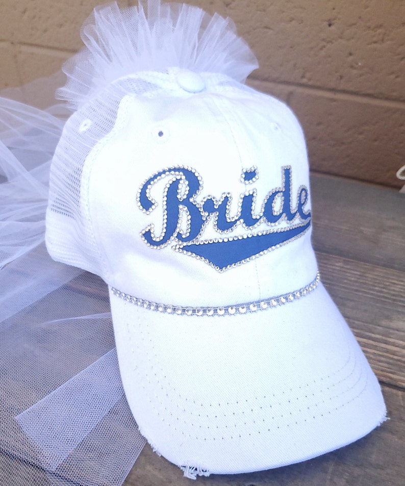 May include: White baseball cap with a tulle veil attached. The cap has a blue and white "Bride" design with rhinestones.