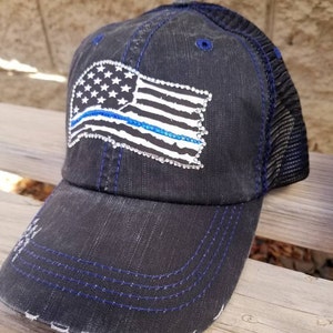 May include: A black and blue baseball cap with a distressed denim look. The cap features a white and blue American flag with a blue line running through it. The flag is covered in rhinestones.