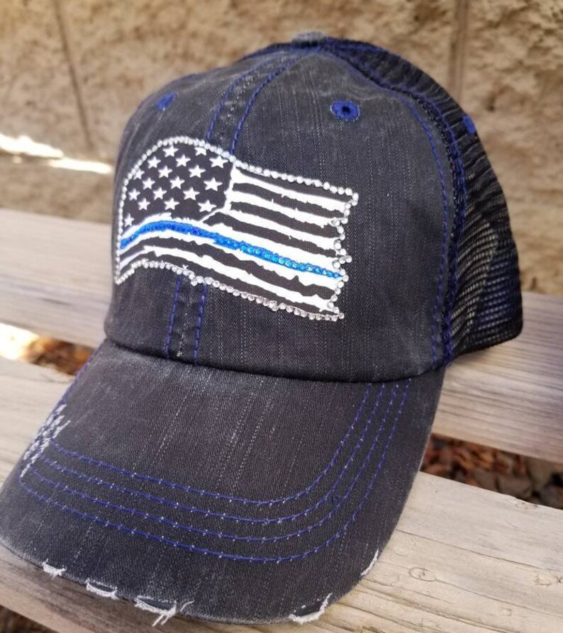 May include: A black and blue baseball cap with a distressed denim look. The cap features a white and blue American flag with a blue line running through it. The flag is covered in rhinestones.