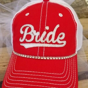 May include: Red and white baseball cap with a white mesh back. The cap has a white rhinestone trim and the word "Bride" in white rhinestones on the front.