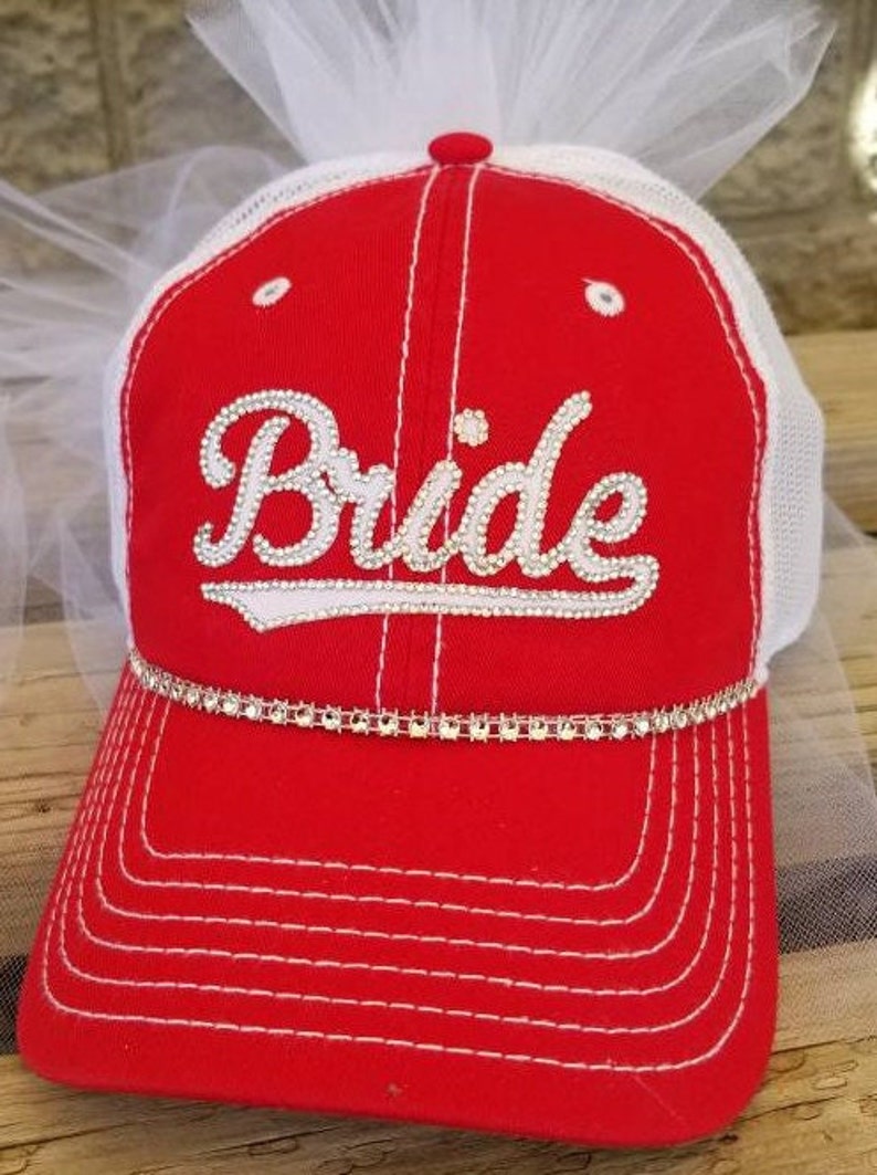 May include: Red and white baseball cap with a white mesh back. The cap has a white rhinestone trim and the word "Bride" in white rhinestones on the front.