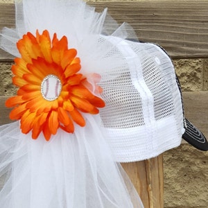 May include: A white baseball cap with a black mesh back and a white tulle veil. An orange flower with a baseball charm is attached to the veil.
