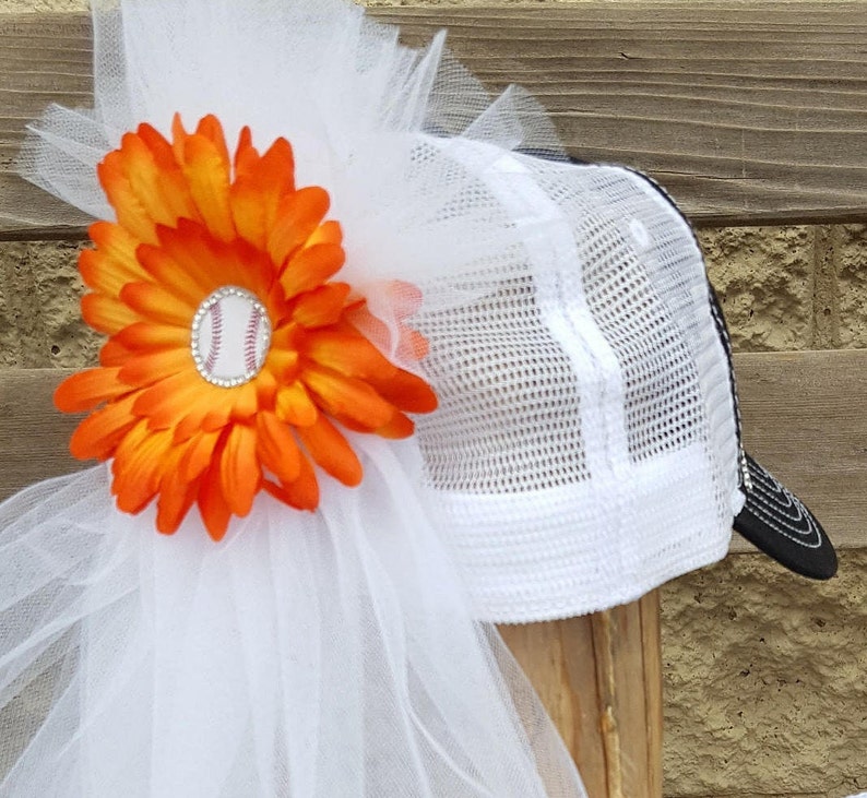 May include: A white baseball cap with a black mesh back and a white tulle veil. An orange flower with a baseball charm is attached to the veil.