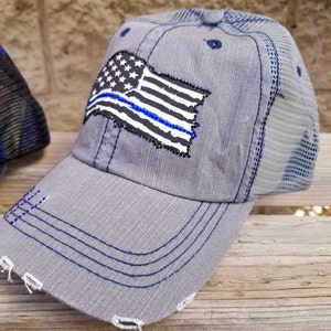 May include: A grey and blue distressed trucker hat with a thin blue line American flag patch. The hat has a mesh back and a frayed brim.