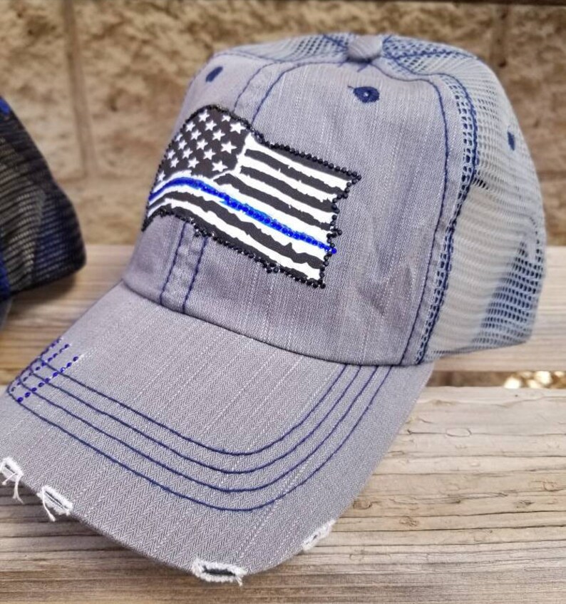 May include: A grey and blue distressed trucker hat with a thin blue line American flag patch. The hat has a mesh back and a frayed brim.