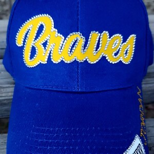 Puede incluir: Gorra de b&eacute;isbol azul con un logotipo amarillo "Braves" con diamantes de imitaci&oacute;n. La gorra tiene un logotipo "Cheer" blanco y negro con un meg&aacute;fono en el lateral.