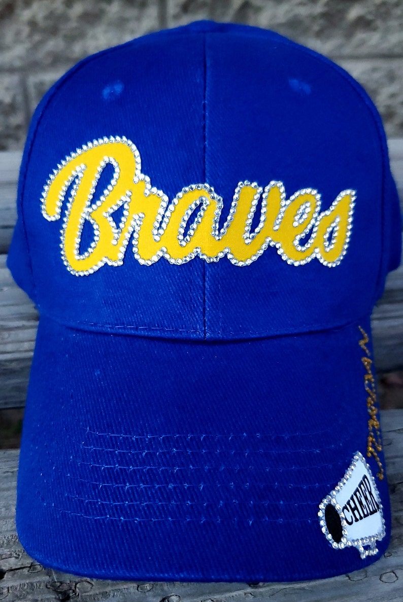 Puede incluir: Gorra de b&eacute;isbol azul con un logotipo amarillo "Braves" con diamantes de imitaci&oacute;n. La gorra tiene un logotipo "Cheer" blanco y negro con un meg&aacute;fono en el lateral.