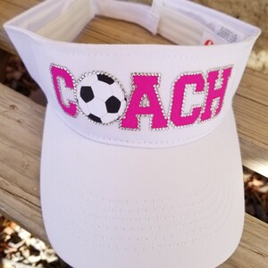 May include: White visor with a pink "COACH" design with rhinestones and a soccer ball in the middle.