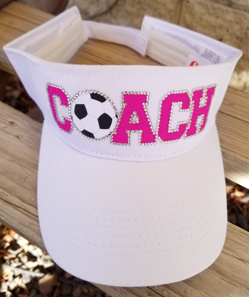 May include: White visor with a pink "COACH" design with rhinestones and a soccer ball in the middle.