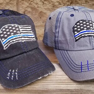 May include: Two distressed baseball caps with a mesh back. Both caps feature a blue line American flag design. One cap is black and the other is grey.