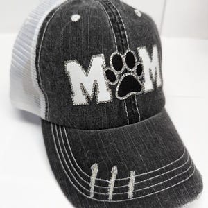 May include: A black and white trucker hat with a distressed denim look. The hat has a white mesh back and a black paw print with rhinestones in the center of the word "MOM" in white rhinestones.