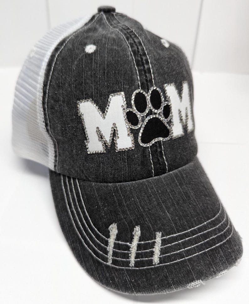 May include: A black and white trucker hat with a distressed denim look. The hat has a white mesh back and a black paw print with rhinestones in the center of the word "MOM" in white rhinestones.