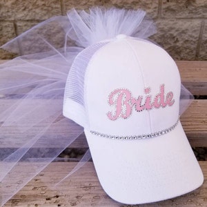 May include: White trucker hat with a pink "Bride" design and rhinestones. The hat has a white tulle veil attached to the top.
