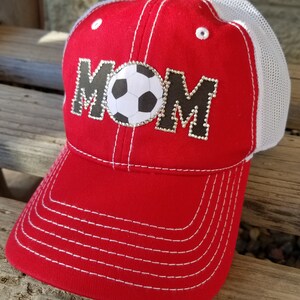 Op de afbeelding: Rode en witte baseballpet met een wit mesh achterpaneel. De voorkant van de pet heeft het woord "MOM" in zwart met strass en een witte voetbal in het midden.