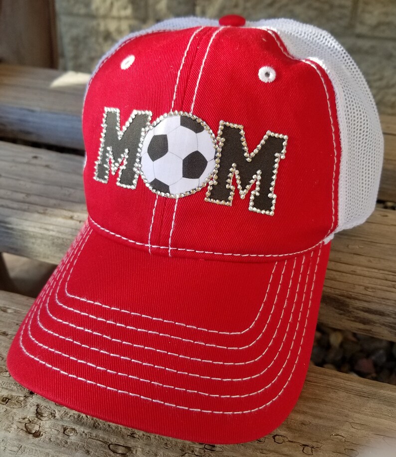 Op de afbeelding: Rode en witte baseballpet met een wit mesh achterpaneel. De voorkant van de pet heeft het woord "MOM" in zwart met strass en een witte voetbal in het midden.