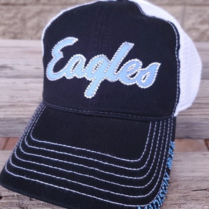 Puede incluir: Gorra de b&eacute;isbol negra y blanca con un logotipo de "Eagles" de diamantes de imitaci&oacute;n azul y blanco. La gorra tiene una parte posterior de malla y una visera blanca.