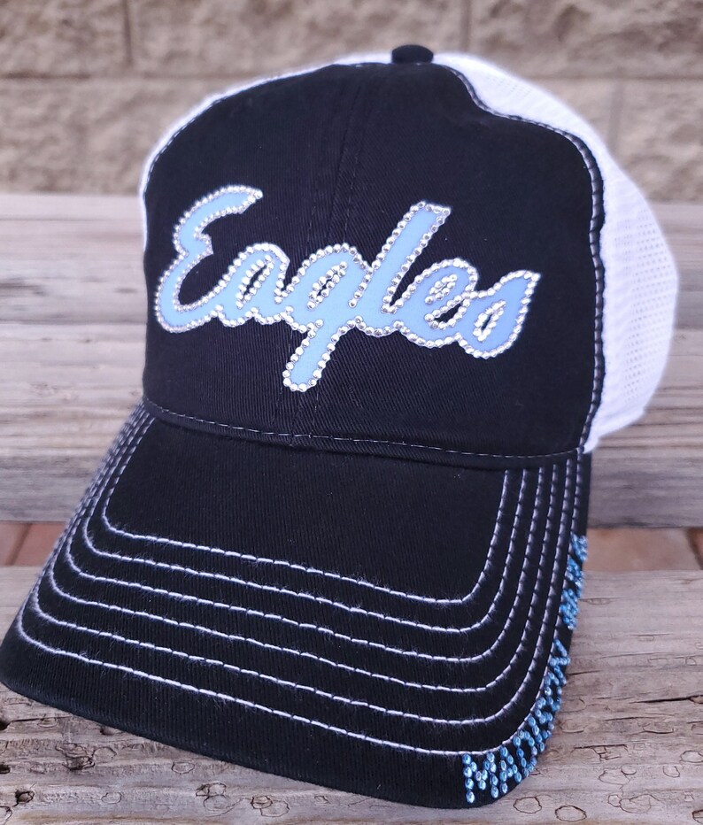 Puede incluir: Gorra de b&eacute;isbol negra y blanca con un logotipo de "Eagles" de diamantes de imitaci&oacute;n azul y blanco. La gorra tiene una parte posterior de malla y una visera blanca.